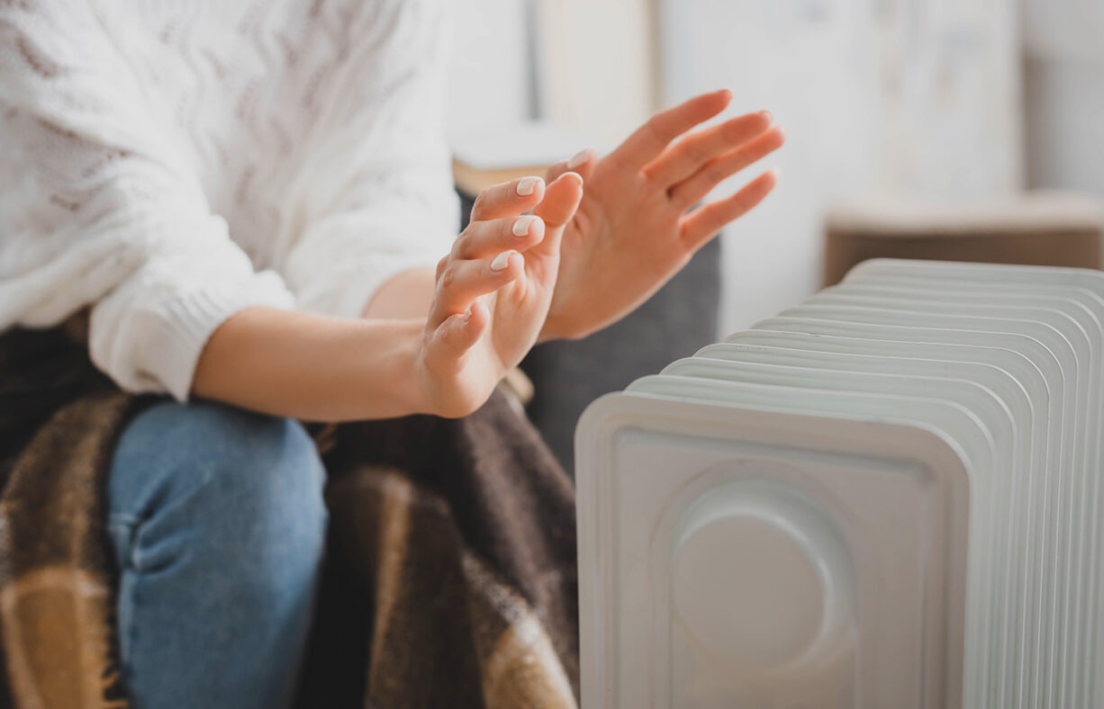 Nainen lämmittelee öljypatterin edessä viltin kanssa. Naisesta näkyy vain kädet ja polvet.